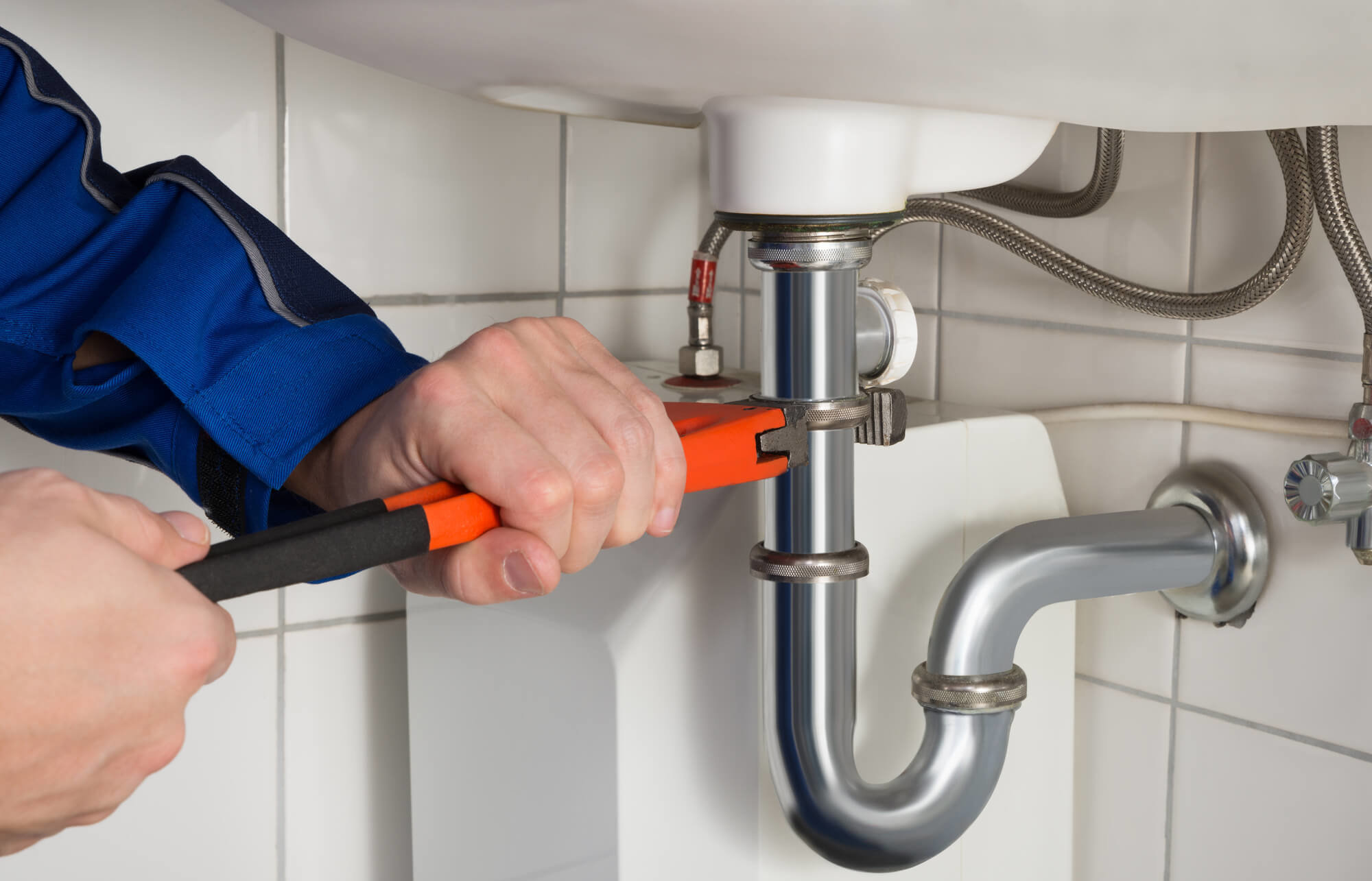 Plumber tightening a chrome sink drain with an orange pipe wrench